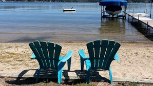 Two blue Adirondack chairs facing a calm lake with a dock and a covered boat.
