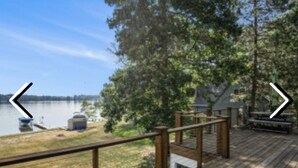 A wooden deck with a railing overlooking a lake, a small dock, and a building with a chimney.