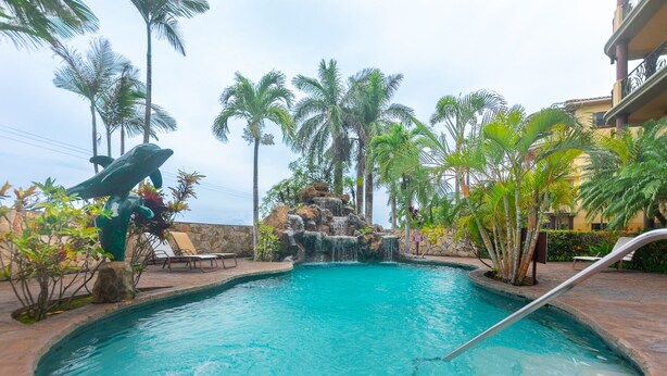A swimming pool with a waterfall feature, surrounded by palm trees and lounge chairs.