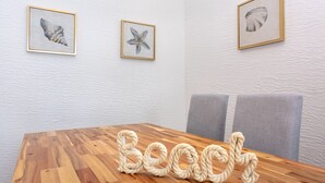 A wooden table with the word "Beach" spelled out in rope letters, flanked by gray chairs. Above the table, there are three framed pictures of seashells on a textured white wall.
