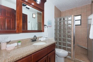 A bathroom with a granite countertop, a white sink, a black faucet, a wooden vanity, a white toilet, and a glass shower enclosure. - Pineapple Villa 442 2 Bedroom Condo by RedAwning (Roatan)