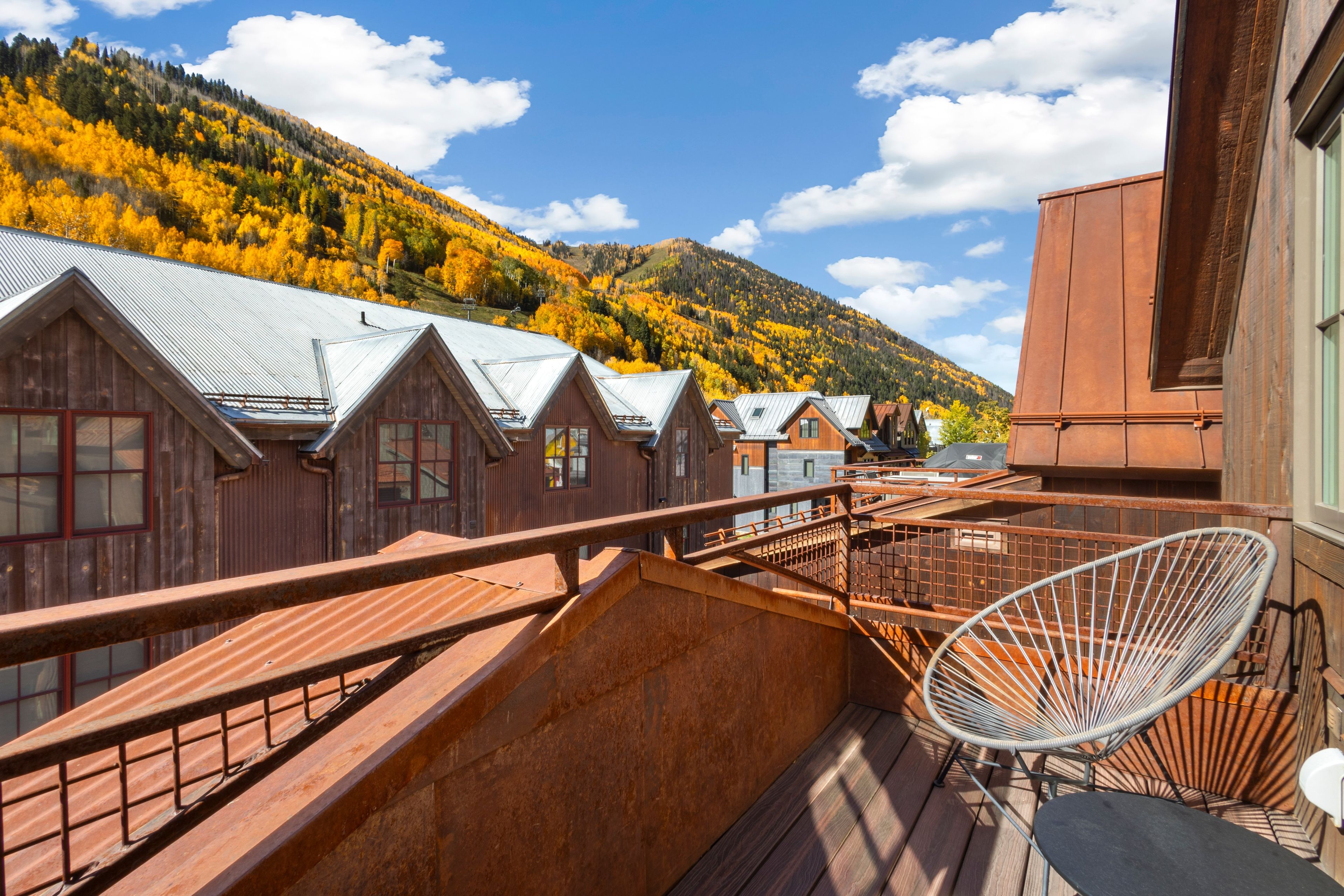 A wooden cabin with a metal roof, a balcony with a chair, and a view of autumn-colored trees in the background.
