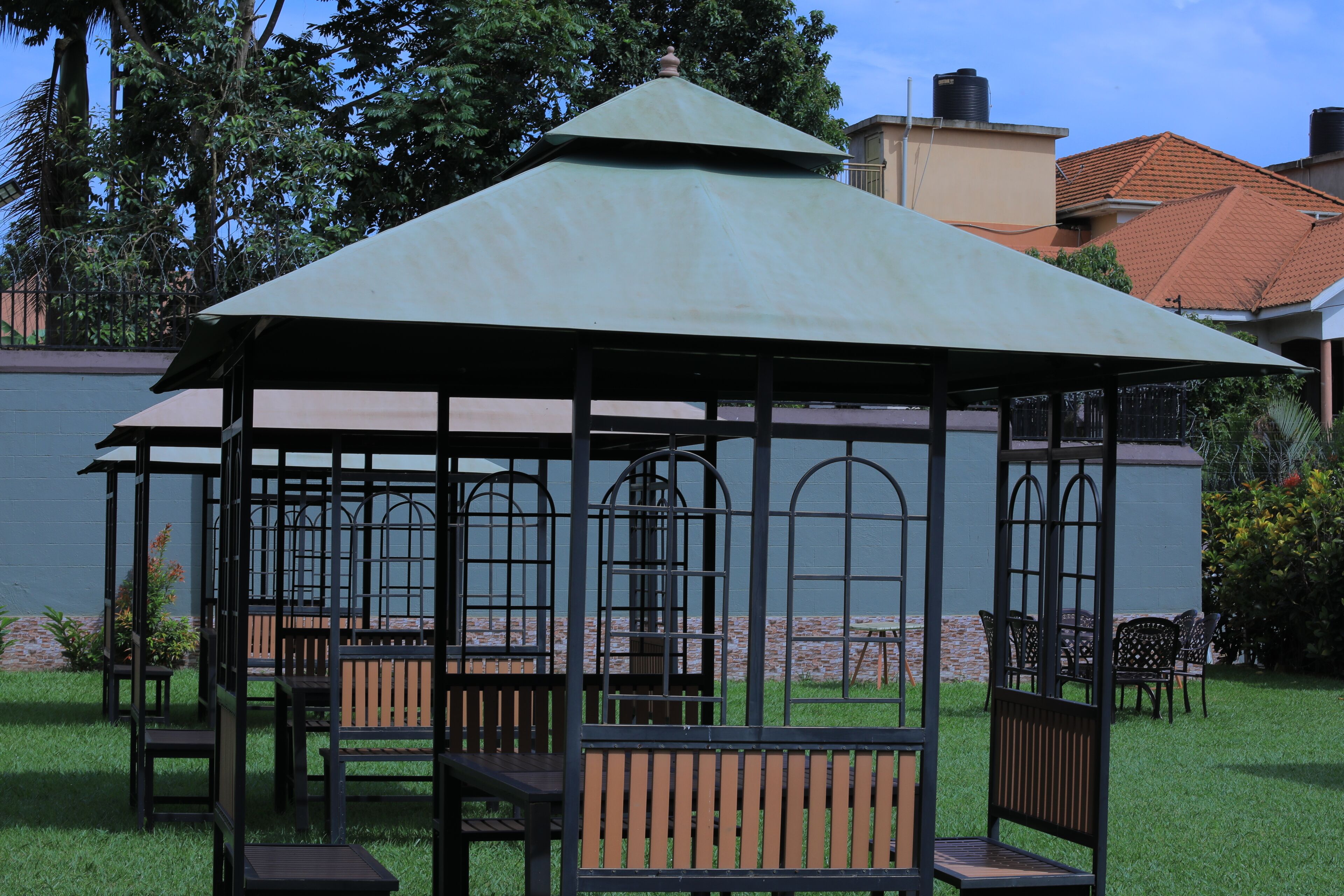 A gazebo with a metal frame and a green roof, surrounded by wooden benches and chairs.