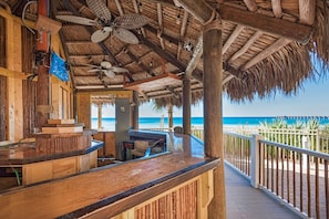 A wooden bar area with a thatched roof, a pool, and a view of the beach.