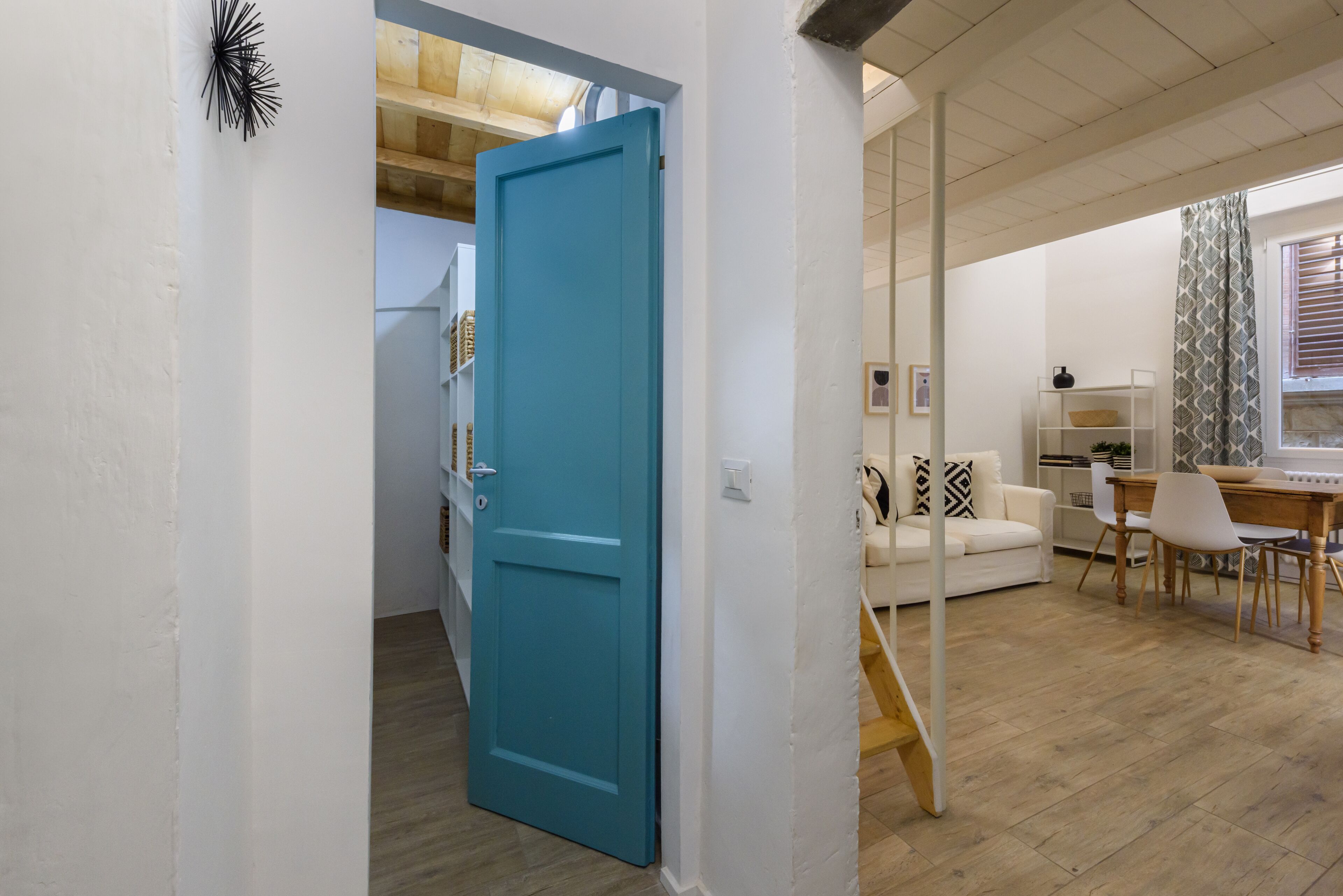A modern interior with a teal door leading to a kitchen area, a dining table with chairs, and a white sofa in a living space.