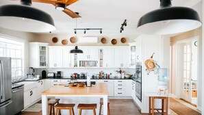 Une cuisine moderne avec des armoires blanches, un îlot central en bois doté de tabourets, un ventilateur de plafond et des luminaires suspendus.