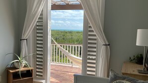 Une chambre avec un grand lit, une table de chevet, une lampe et une vue sur un balcon équipé d’un hamac.
