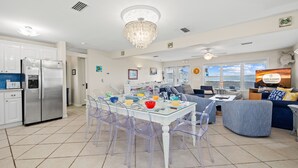Una cocina moderna con electrodomésticos de acero inoxidable, un comedor con sillas traslúcidas y una mesa de vidrio, y una sala de estar con un sofá azul y un televisor que muestra 'Cape & Coast'.