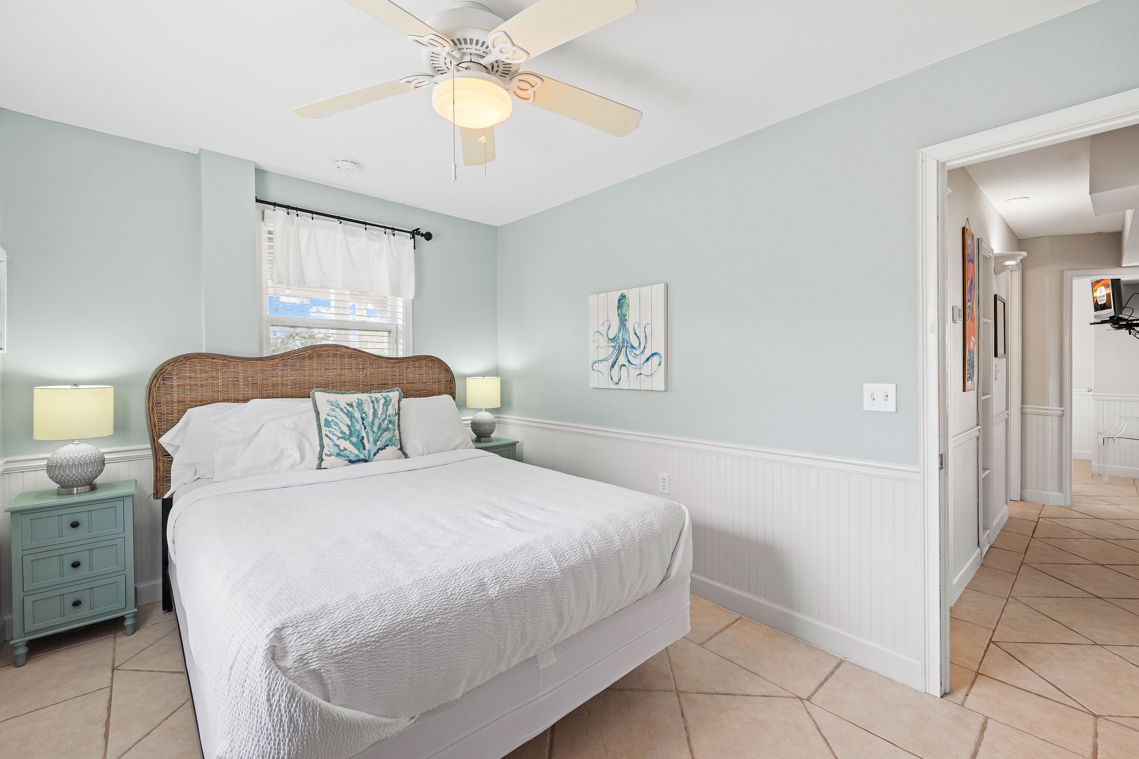A bedroom with a bed, nightstands, a ceiling fan, a window with curtains, and a wall-mounted artwork.