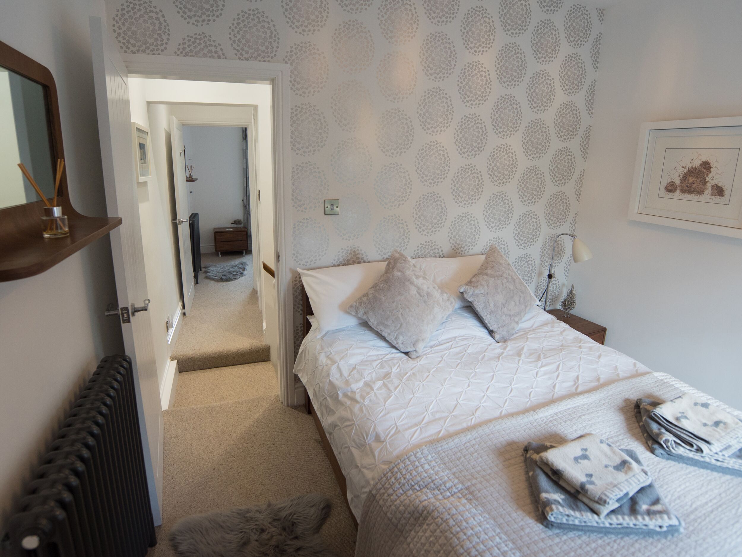 A bedroom with a bed, a nightstand, a lamp, a radiator, and a door leading to another room.