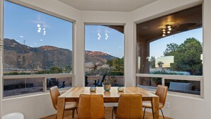 Comedor con mesa de madera y cuatro sillas, ventanas amplias con vistas a las montañas y un ventilador de techo.
