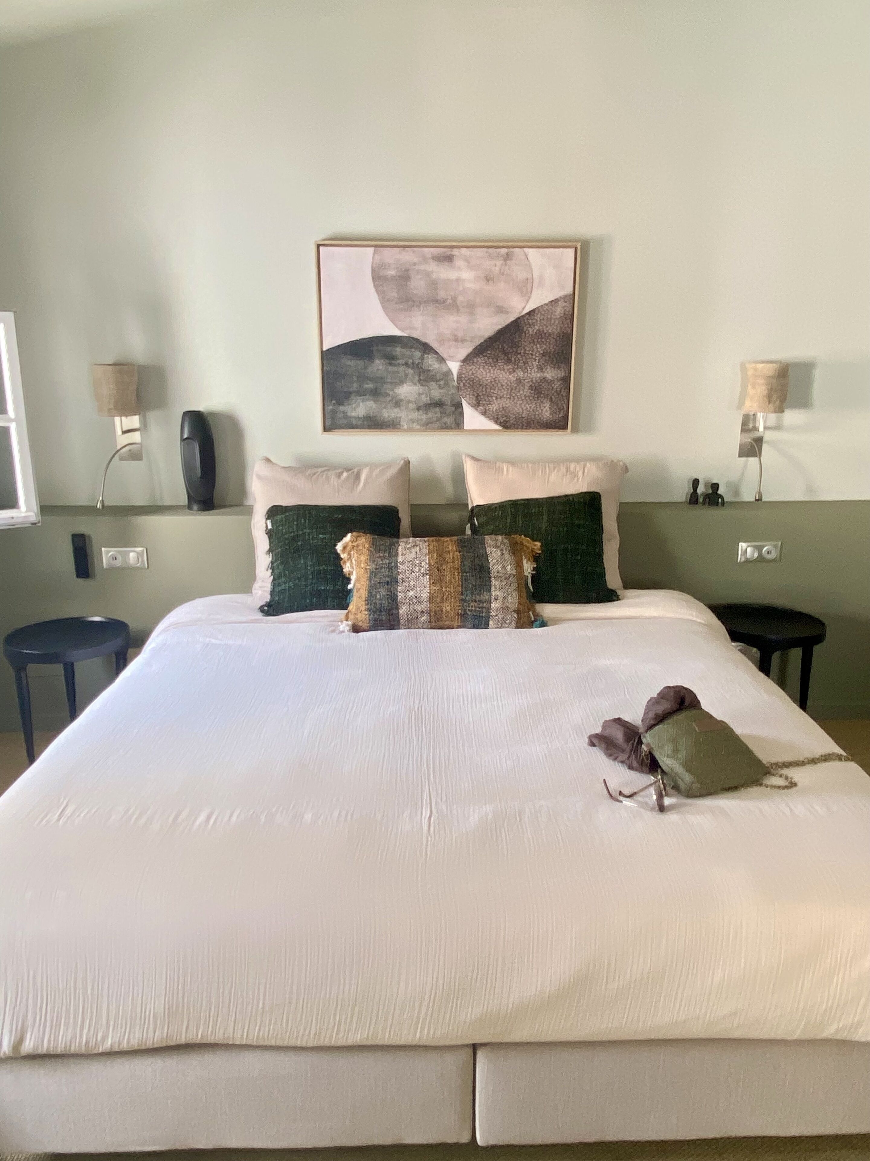 A neatly made bed with a white comforter and several pillows, a wall-mounted abstract painting, bedside lamps, and a small table with a lamp.