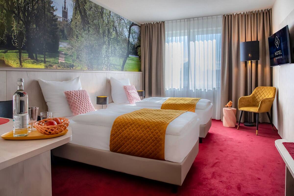 Ein Hotelzimmer mit zwei Betten, einem großen Fenster mit Vorhängen, einem roten Teppich, einem Schreibtisch mit Flasche und Gläsern sowie einem Baumgemälde.