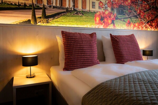Une chambre d’hôtel avec un grand lit, deux coussins rouges à motifs, une couette blanche et une vue sur un jardin aménagé, avec un bâtiment en arrière-plan.