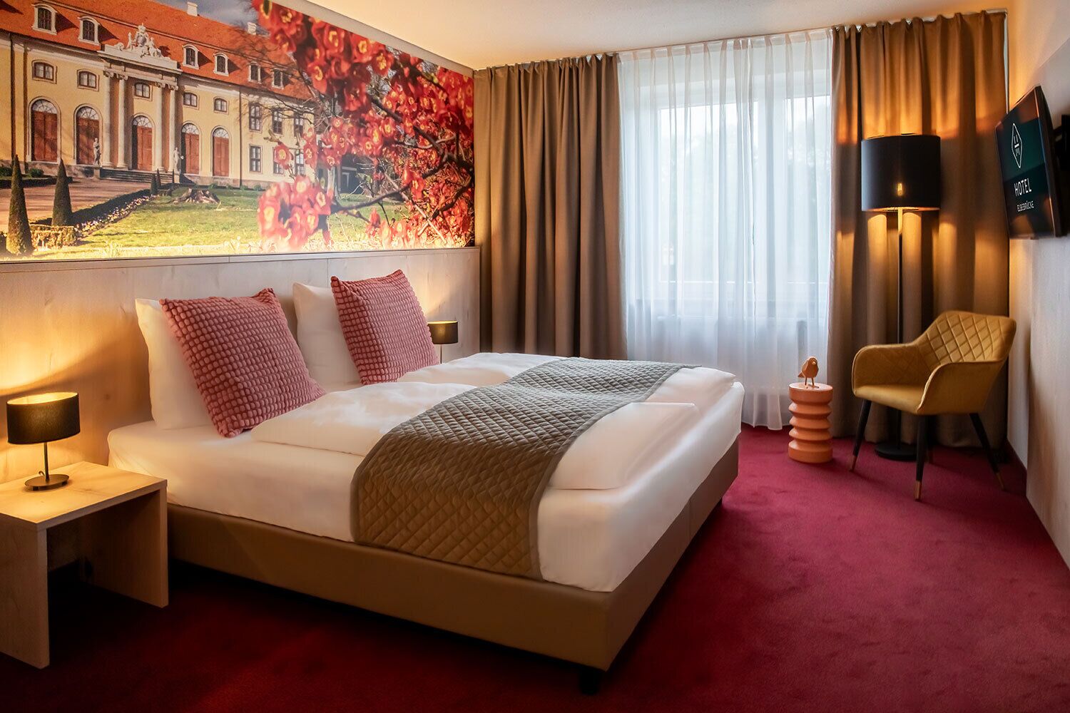 A hotel room with a large bed, a nightstand, a lamp, a chair, and a painting on the wall.