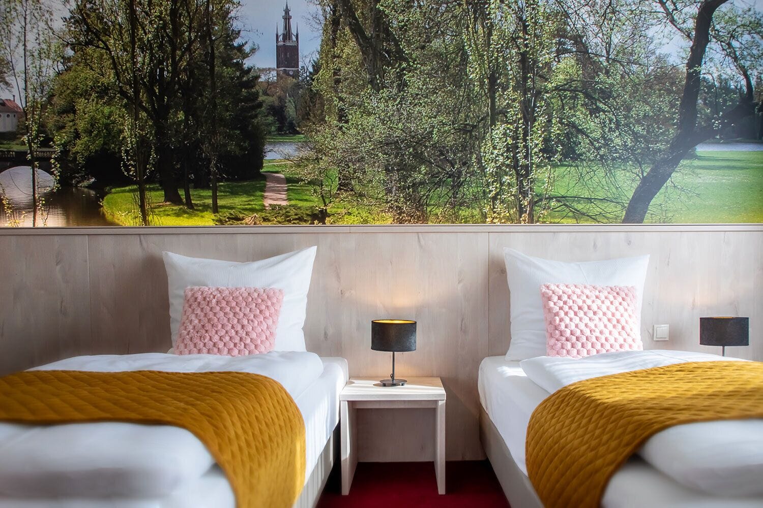 Ein Hotelzimmer mit zwei Betten, jeweils mit einer weißen Daunendecke und einer gelben Tagesdecke. Zwischen den Betten befindet sich ein Nachttisch mit einer Lampe. Das Zimmer verfügt über ein großes Fenster mit Blick auf einen Park und ein Gebäude in der Ferne.
