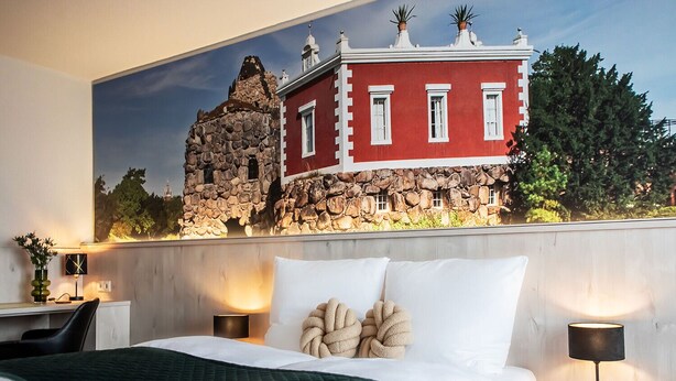 Quarto de hotel com uma cama, duas mesas de cabeceira, uma escrivaninha e um grande mural de um edifício vermelho com paredes de pedra.
