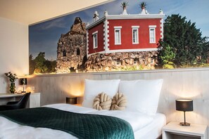 Une chambre d’hôtel comprenant un lit, deux tables de chevet, un bureau et une grande fresque murale représentant un bâtiment rouge aux murs de pierre.