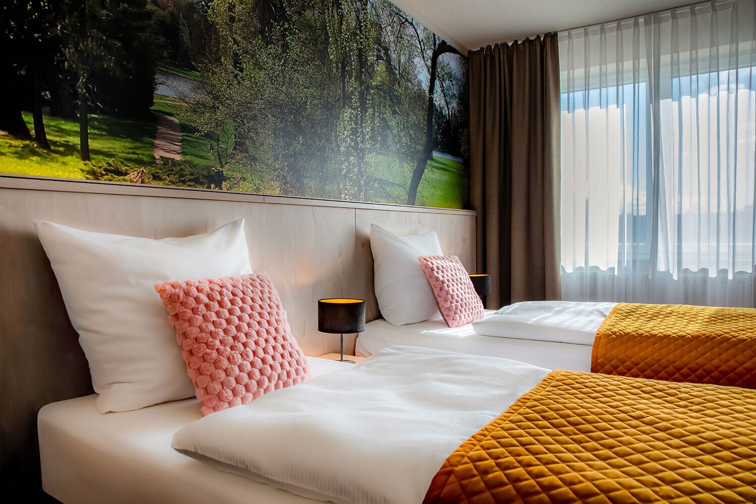 A hotel room with two beds, a large window with curtains, a wall mural, and a bedside table with a lamp.