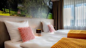 A hotel room with two beds, a large window with curtains, a wall mural, and a bedside table with a lamp.