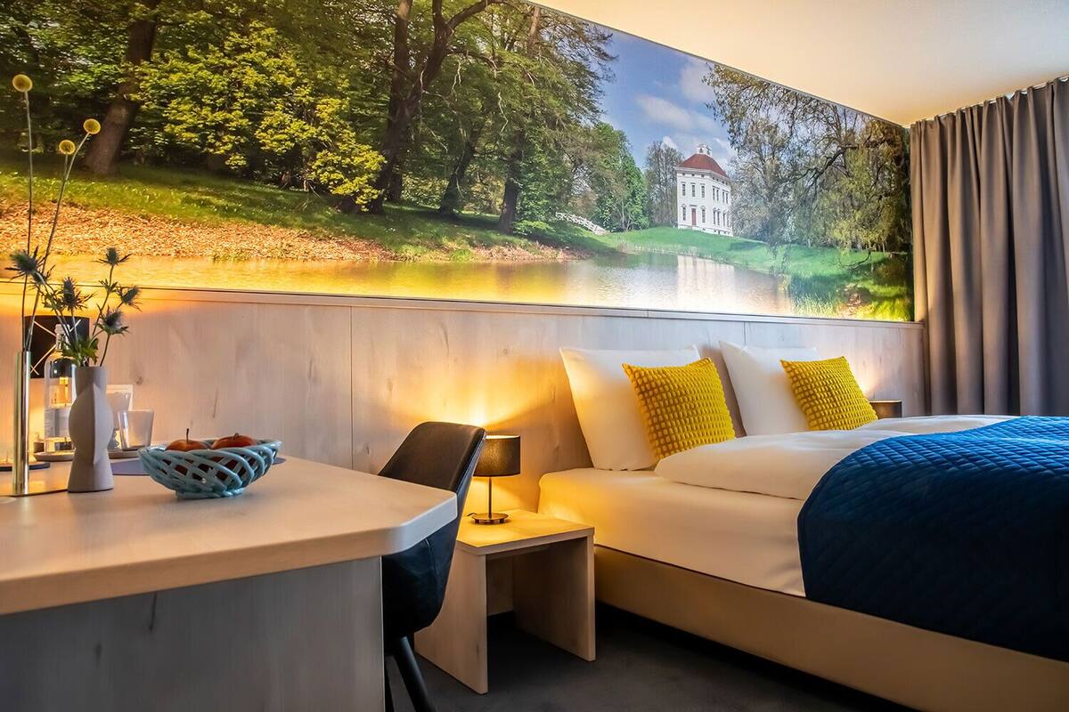 Ein Hotelzimmer mit Bett, Nachttisch, Lampe und einem Wandbild einer Landschaft mit einem Gebäude.