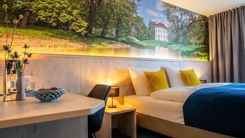 Habitación de hotel con cama, mesita de noche, lámpara y un mural con un paisaje que incluye un edificio.
