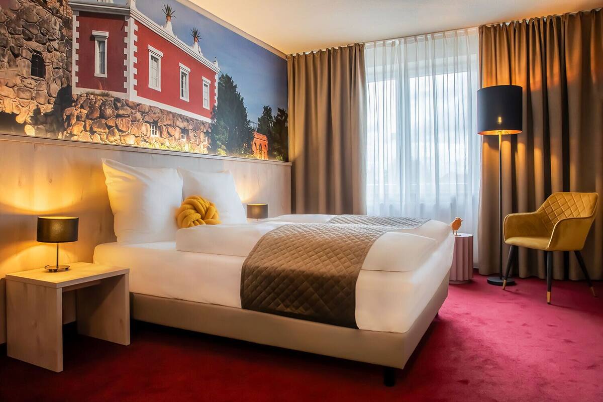 Ein Hotelzimmer mit einem großen Bett, einem Nachttisch mit Lampe, einem Sessel und einem Wandgemälde.