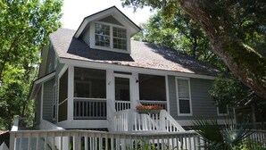 Une maison à deux étages avec un porche, une petite terrasse et un grand arbre en arrière-plan.