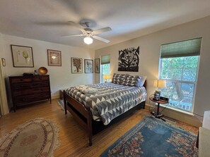 Une chambre à coucher comprenant un lit, une commode, un ventilateur de plafond, des œuvres d’art et une fenêtre donnant sur un paysage agréable.