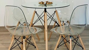 A dining area with a glass table and transparent chairs, a decorative wall piece, and a small bowl of snacks on the table.