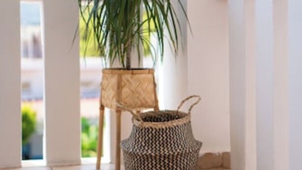 Una cesta tejida con asa, colocada sobre un suelo de baldosas junto a una silla de mimbre.