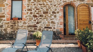 Un bâtiment en pierre avec deux fenêtres et une porte, deux fauteuils et des plantes en pot.