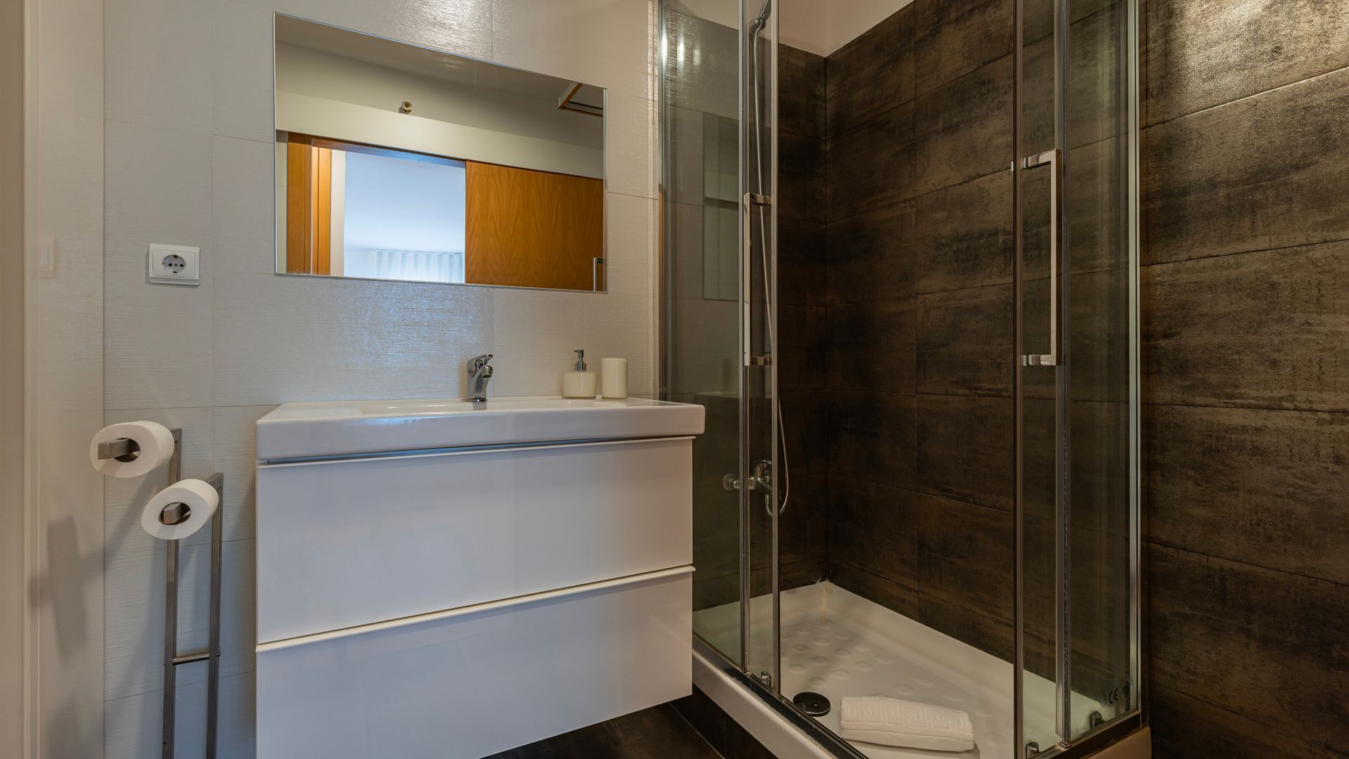 Een moderne badkamer met een douchecabine van glas, een wit wastafelmeubel met spiegel en een handdoek op de vloer.