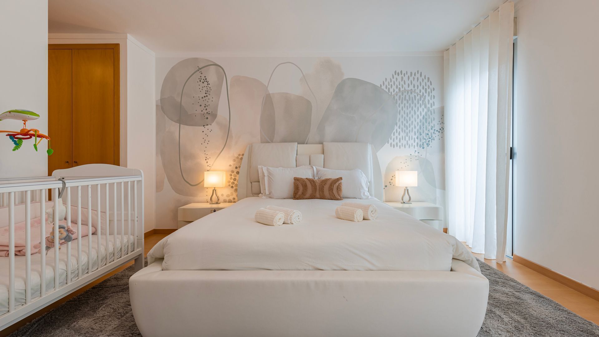 A modern bedroom with a large bed, a crib, and abstract wall art.