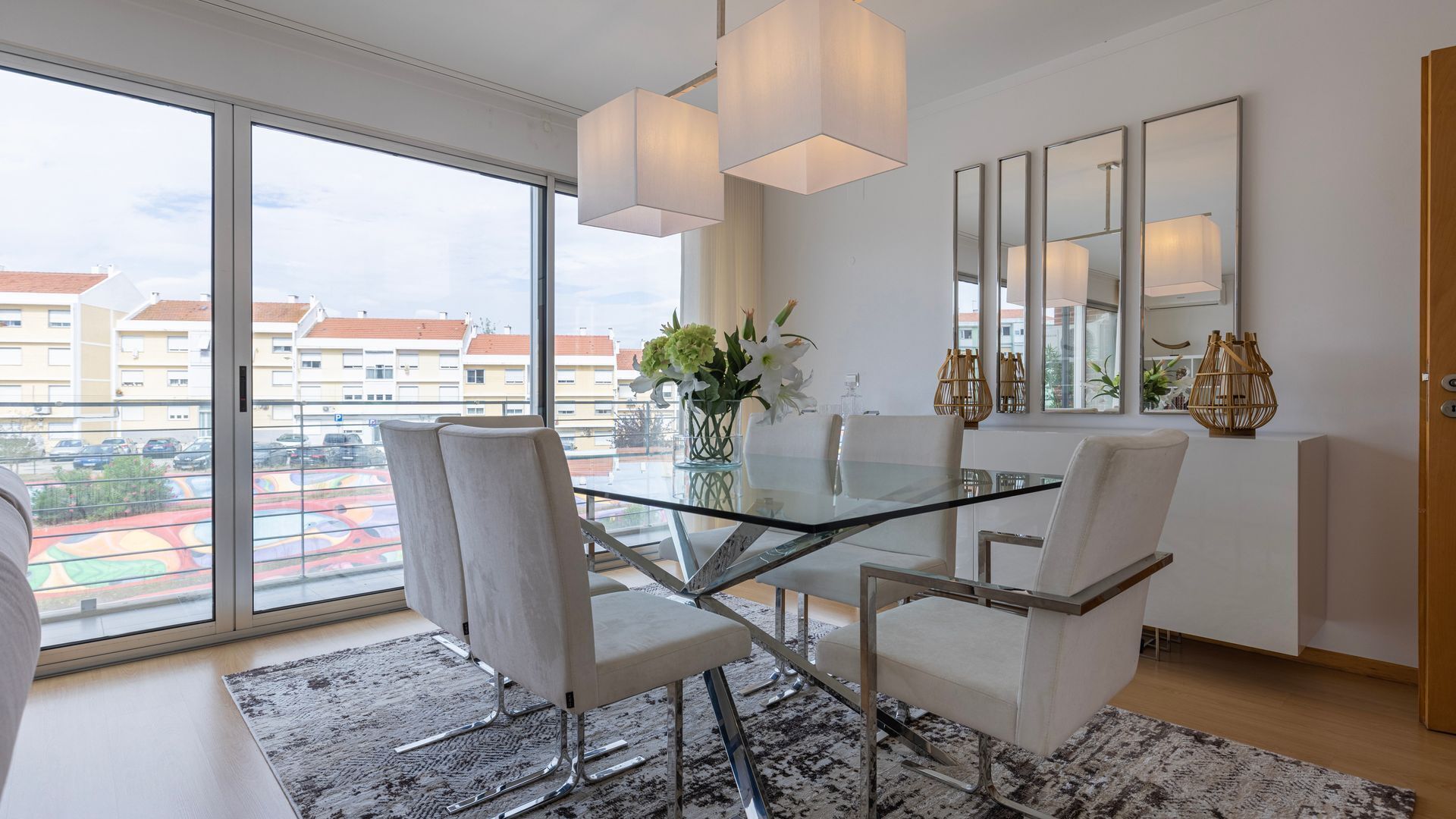 Un comedor moderno con una mesa de vidrio, sillas blancas y vistas a una zona residencial a través de amplios ventanales.