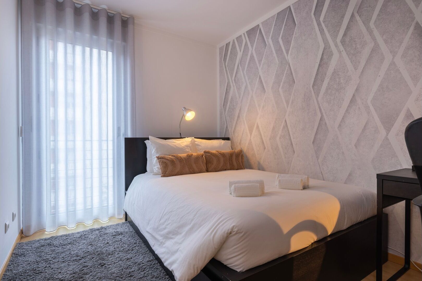 Une chambre d’hôtel moderne avec un lit, une table de chevet, une lampe et un mur orné d’un motif géométrique.