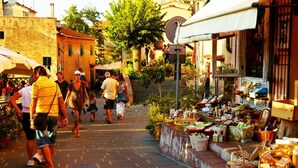 Uma rua de paralelepípedos com pessoas caminhando, um quiosque de mercado com diversos produtos e edifícios com varandas.