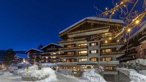 A multi-story hotel with balconies, a snow-covered entrance, and illuminated string lights. - Seven Alpina Boutique Hotel (Klosters-Serneus)