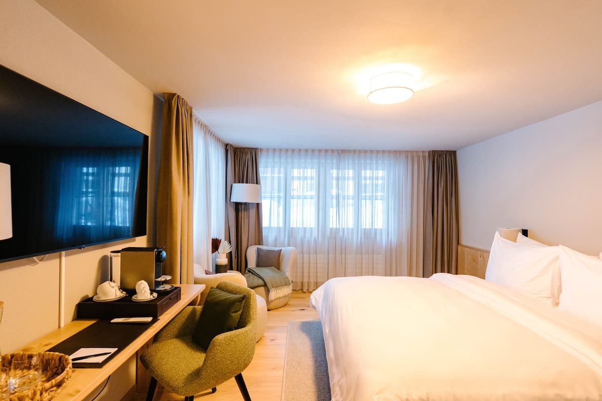 Habitación de hotel con cama, escritorio, silla, televisor y ventana con cortinas.