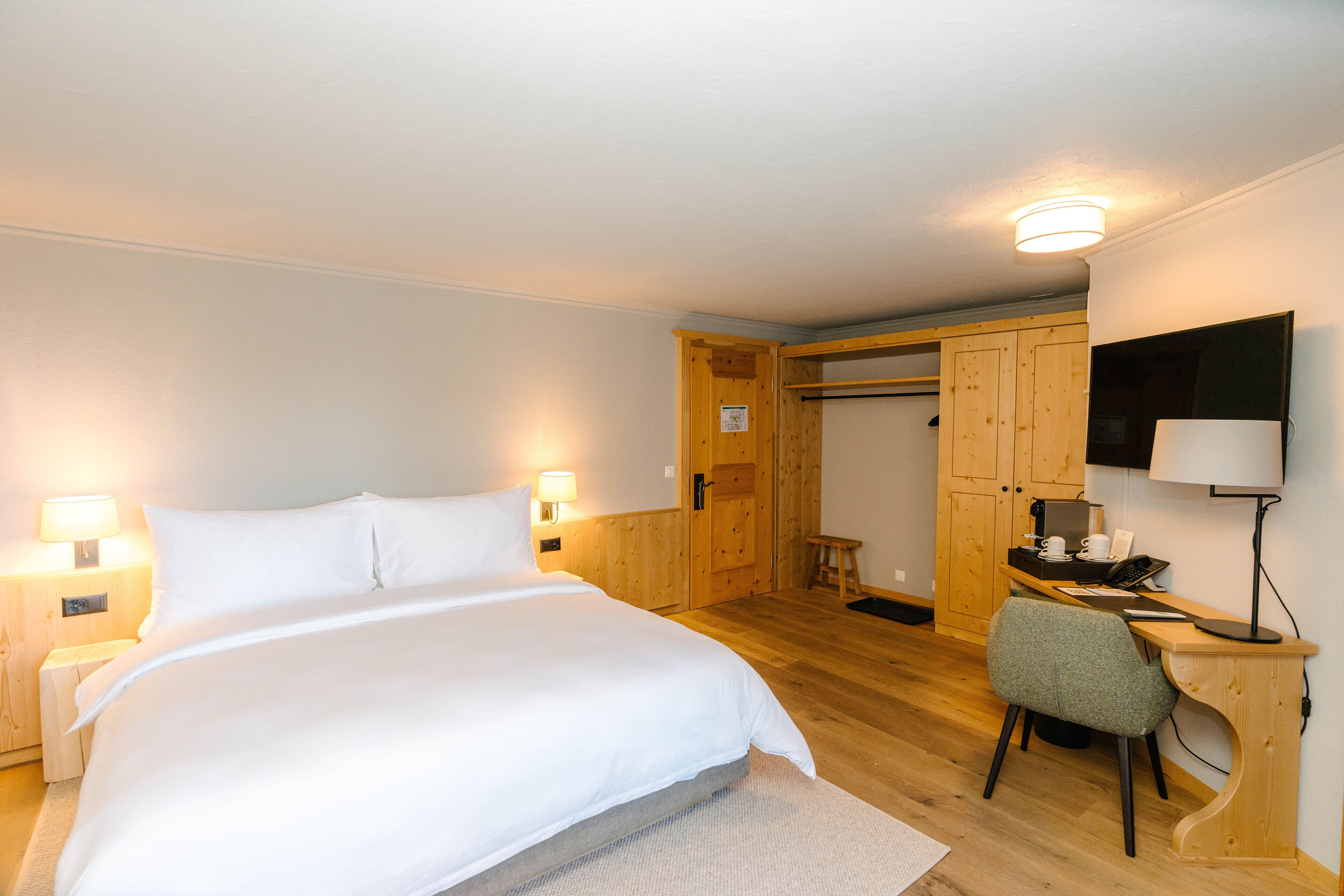 A bedroom with a large bed, bedside lamps, a wooden desk with a chair, a television, and a wardrobe.