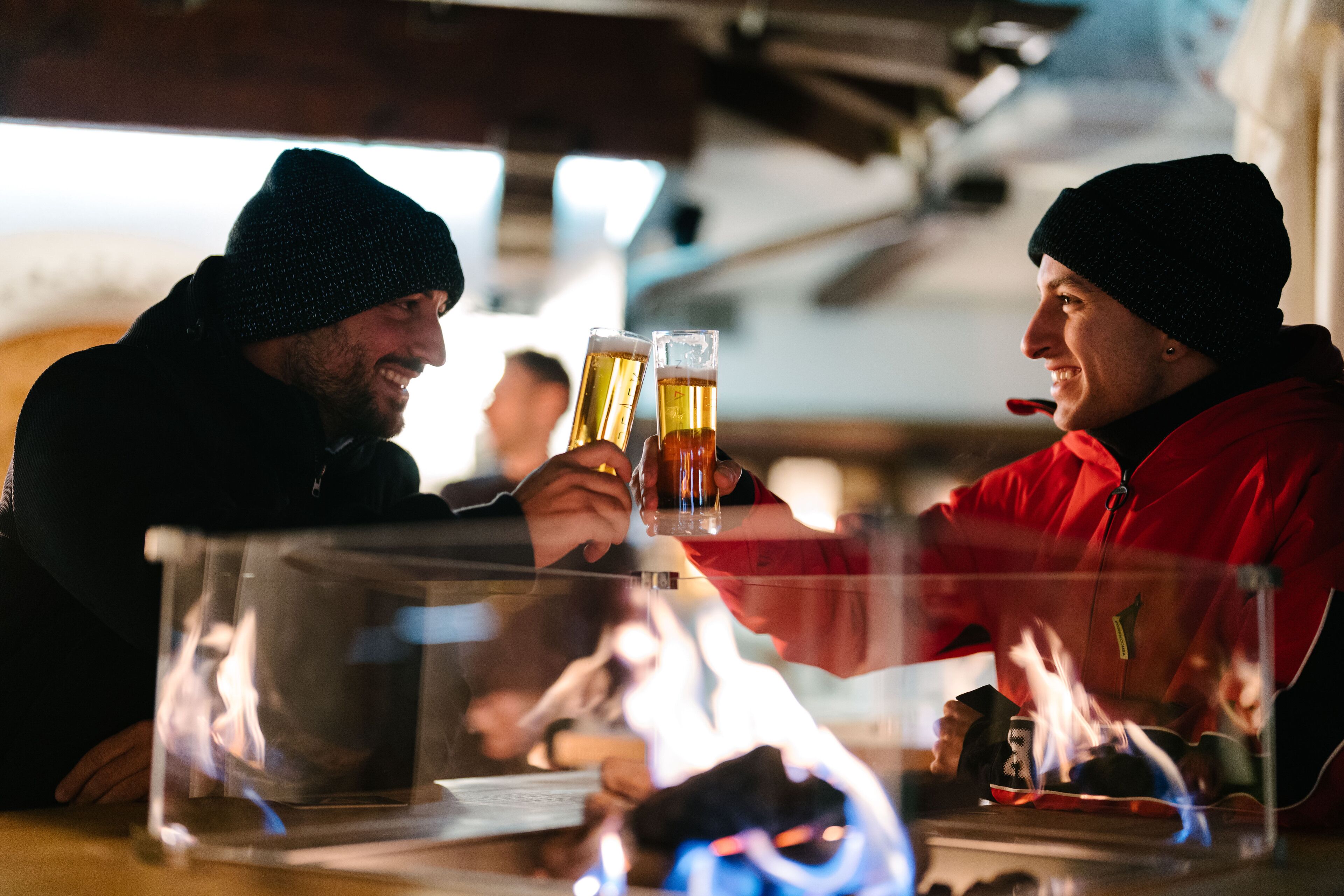 Dos personas brindando con cervezas en un bar con un separador de vidrio transparente.