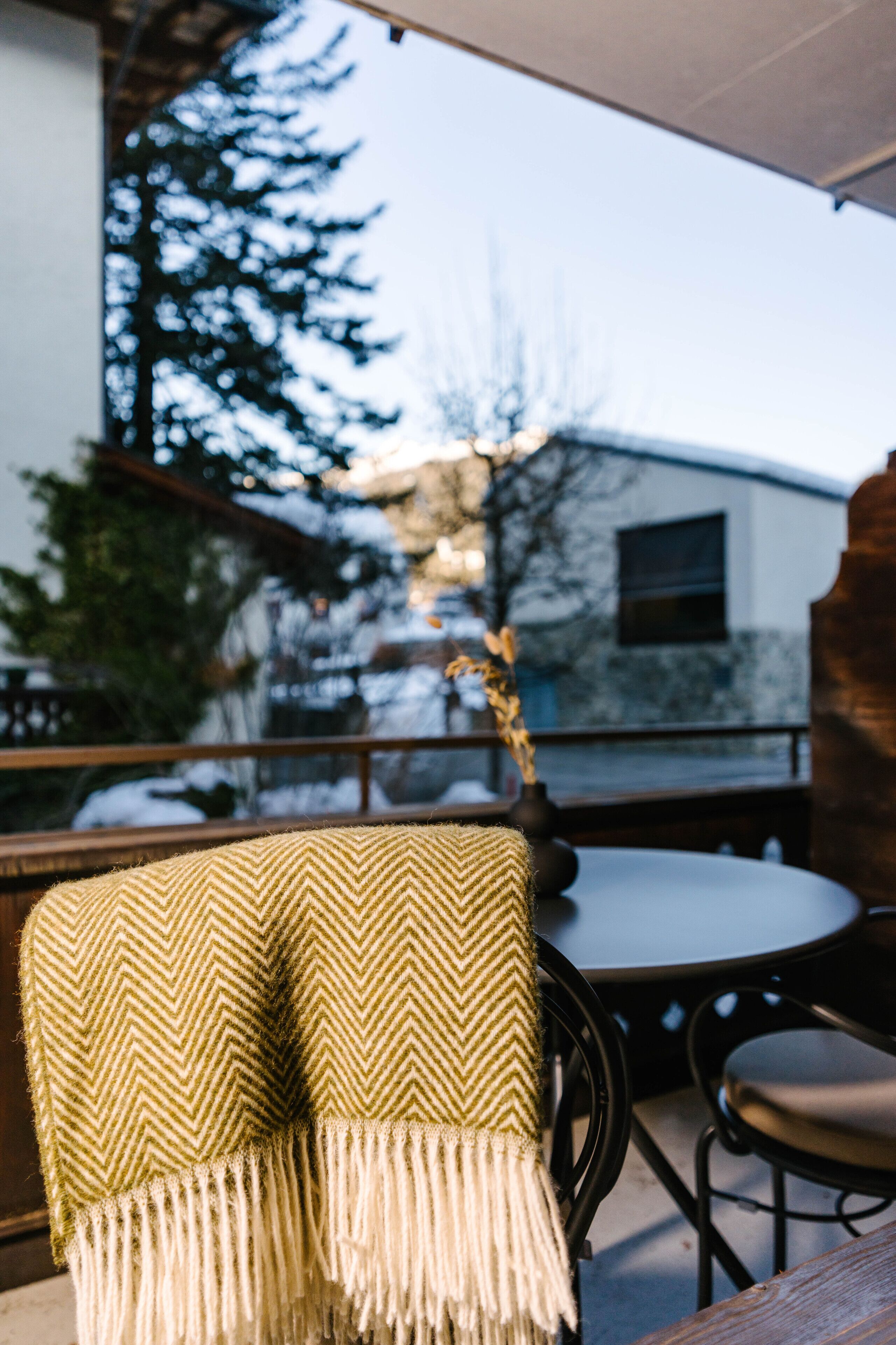 Cómoda zona de estar al aire libre con una mesa y sillas de madera, una manta estampada verde y blanca sobre una silla, y vistas de un edificio y árboles al fondo.