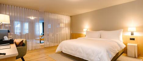 A modern hotel room with a large bed, a desk with a chair, and a view of the city through sheer curtains.