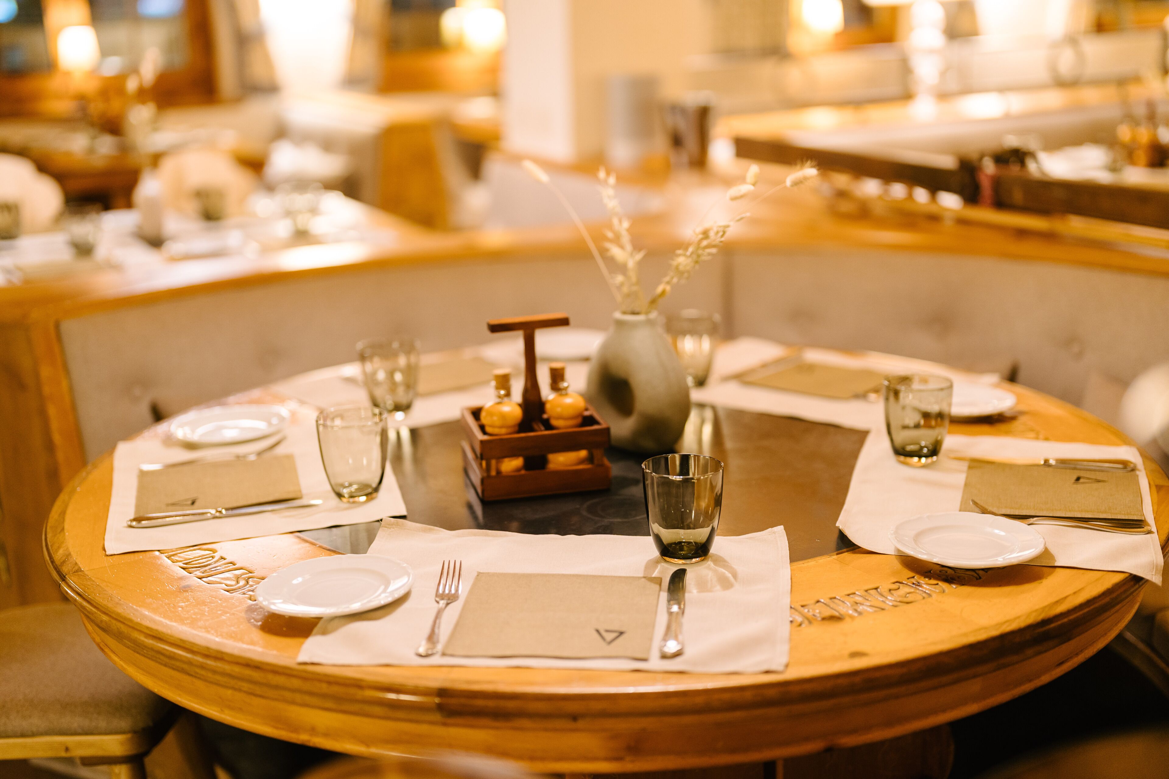 Een ronde eettafel gedekt voor vier personen, met witte borden, bestek en glazen, een vaas met gedroogde bloemen en een klein houten doosje.