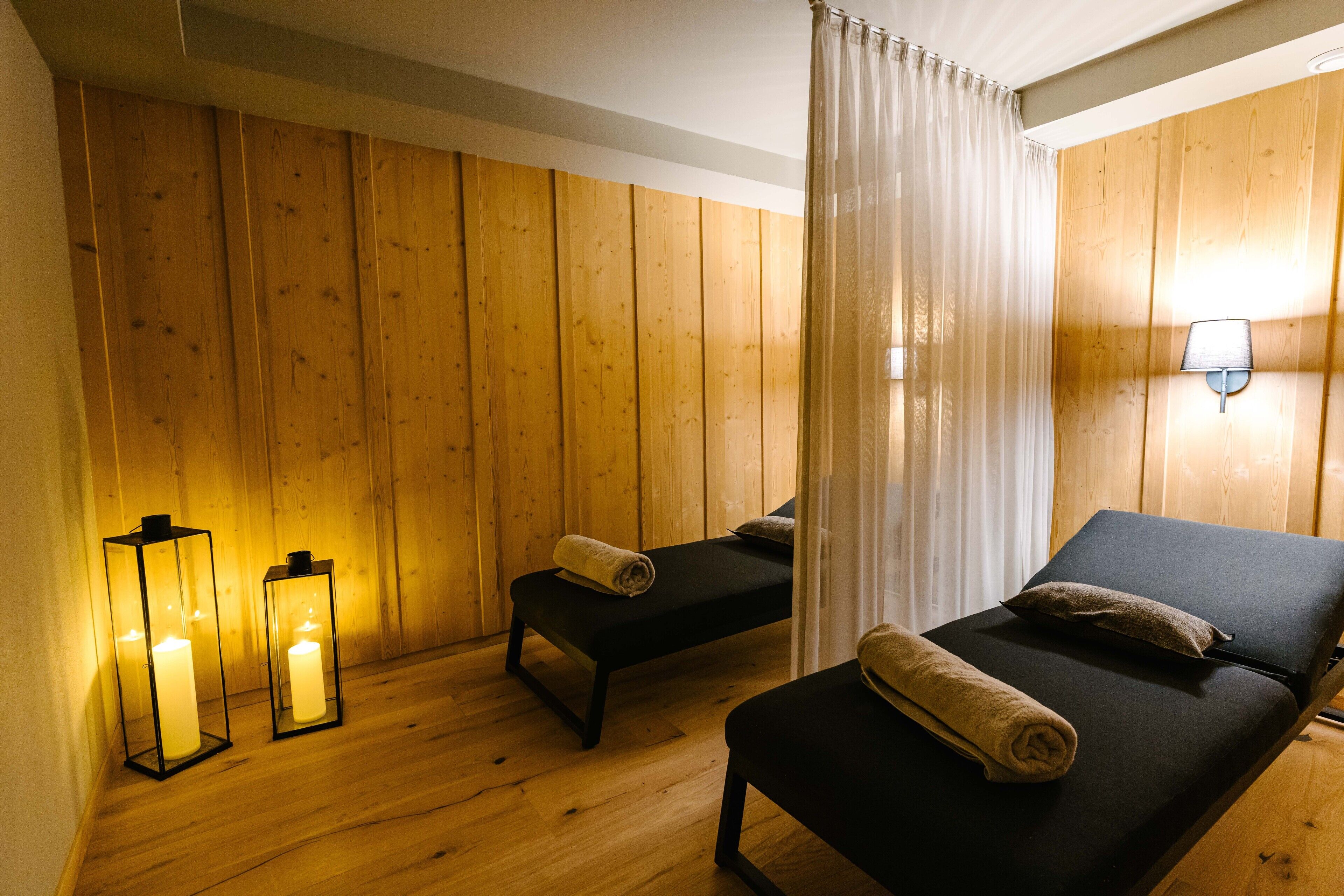 Ein Zimmer mit Holzwänden, einem Bett mit Kopfkissen und zusammengelegtem Handtuch, einer Bank mit Handtuch sowie zwei beleuchteten Laternen.