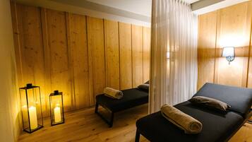 Ein Zimmer mit Holzwänden, einem Bett mit Kopfkissen und zusammengelegtem Handtuch, einer Bank mit Handtuch sowie zwei beleuchteten Laternen.