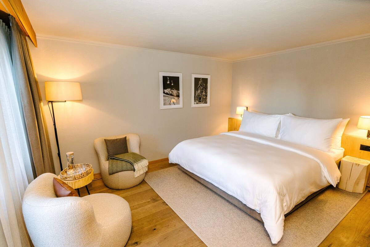 Habitación de hotel con una cama grande, dos mesitas de noche con lámparas, una mesita redonda con lámpara, una silla y dos cuadros en la pared.