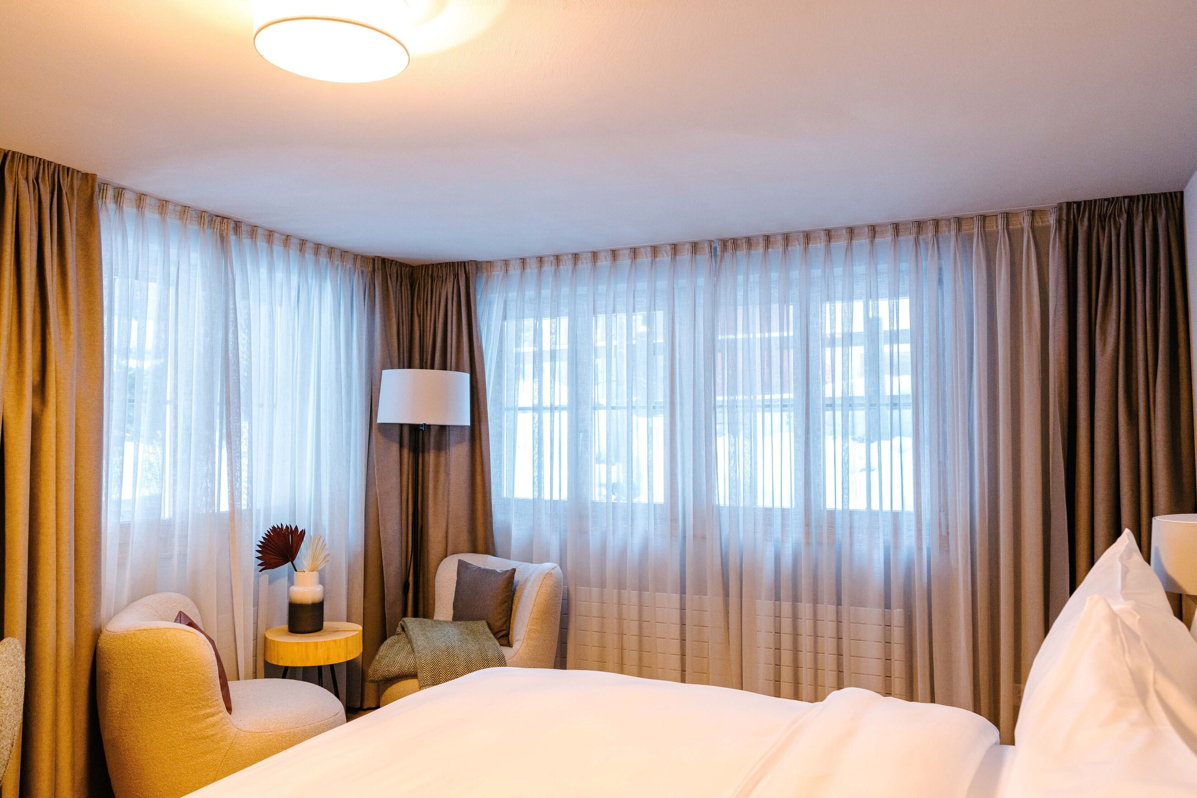 A hotel room with a bed, two chairs, a small table with a lamp, and sheer curtains.