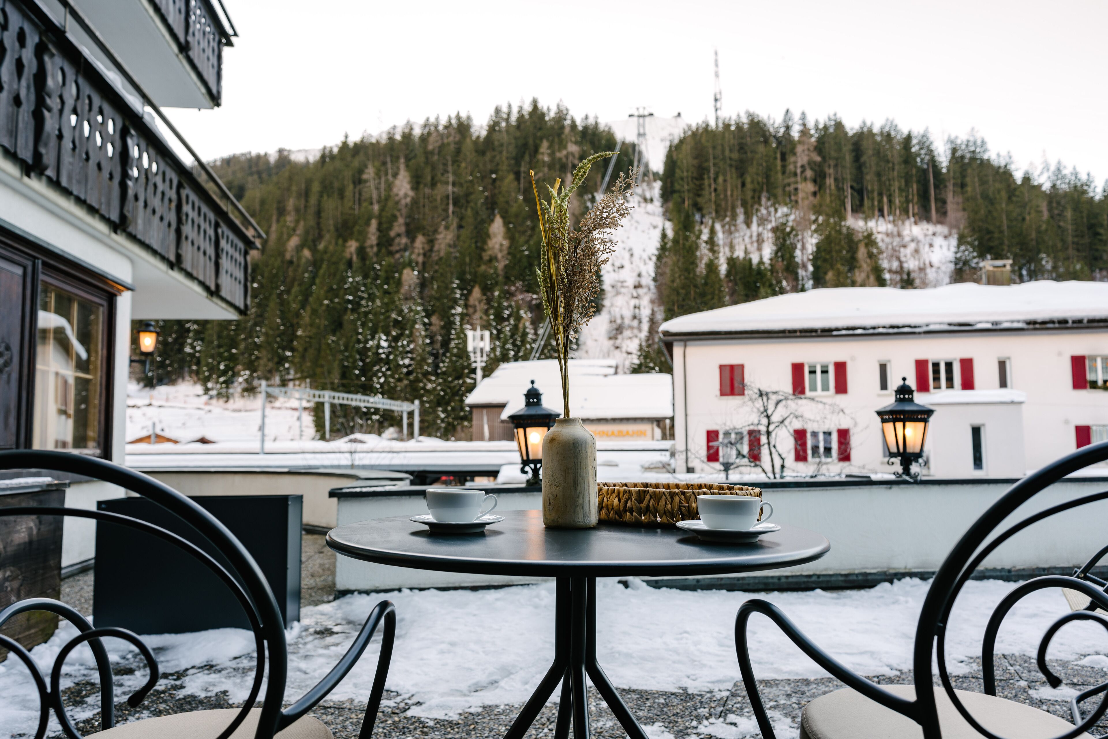 Une table ronde d’extérieur avec des chaises, un vase et deux tasses, le tout posé sur une terrasse enneigée.