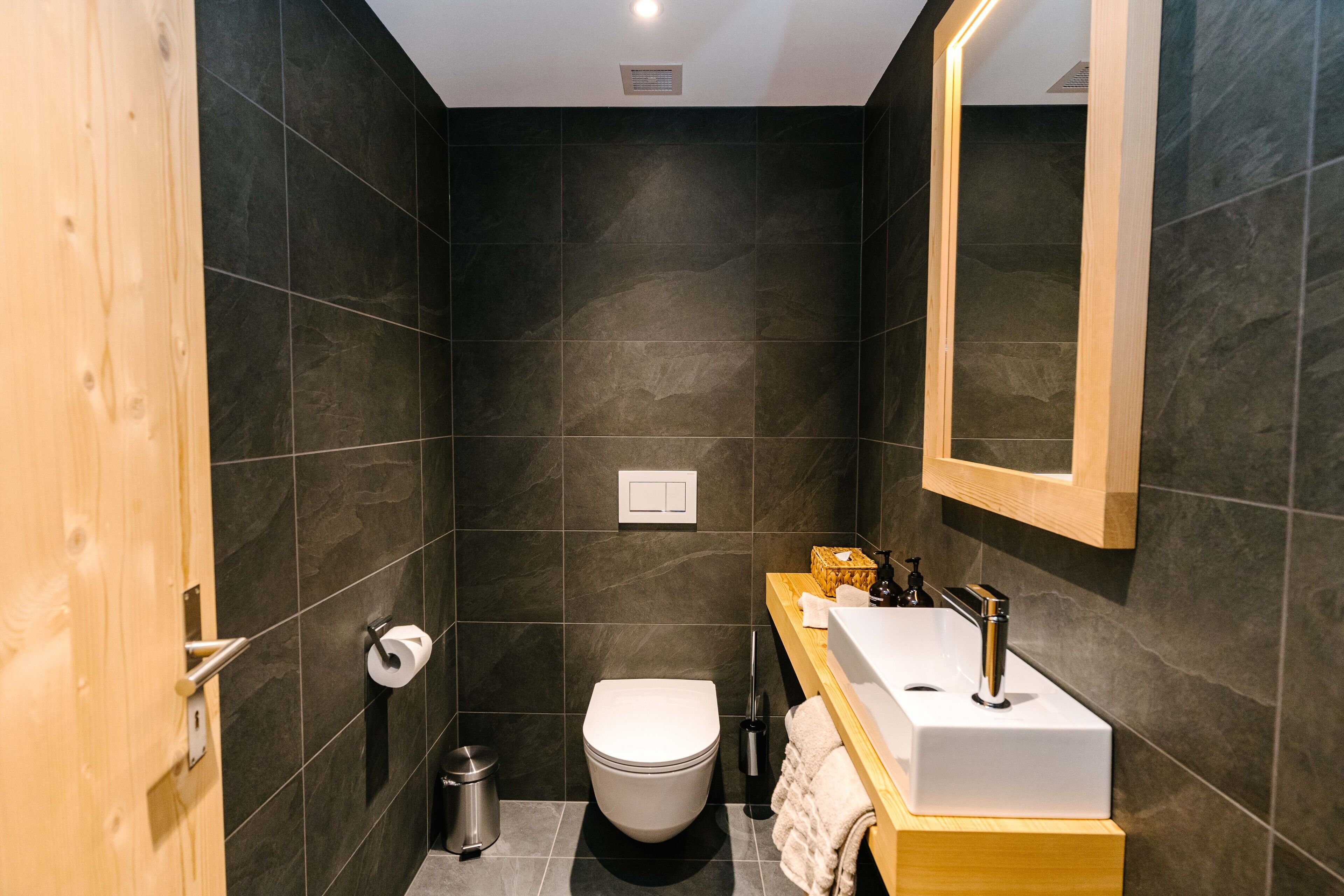 A modern bathroom with dark tiles, a white toilet, a wooden vanity with a sink, and a mirror.